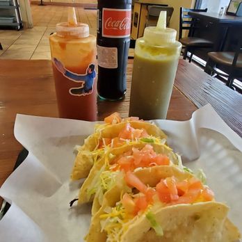 Juanito's Tacos - 4 tacos, red hot sauce bottle, green hot sauce bottle, and a glass bottle coke cola inside the dining area. - Mexican Near Me - Highland, California