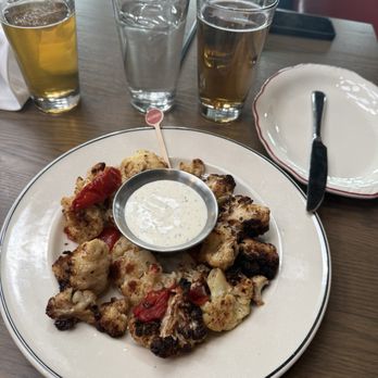 JT Commons - Parmesan cauliflower - A round plate contains roasted cauliflower florets with some charred edges, garnished with red peppers, and a small metal cup of creamy dipping sauce in the center. The plate is on a wooden table with four glasses of beer and water behind it. An empty decorative plate with a fork is also visible. The setting appears to be a casual dining restaurant. - Pizza Near Me - Middletown, Rhode Island