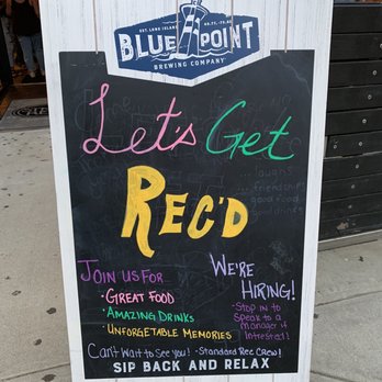 Standard Rec - A chalkboard sign outside Blue Point Brewing Company displays promotional messages in colorful handwriting. It invites customers to enjoy great food, amazing drinks, and unforgettable memories, and mentions that the business is hiring. The sign encourages visitors to stop in and speak to a manager if interested, and ends with a message to relax and sip back. - Bars Near Me - Patchogue, New York