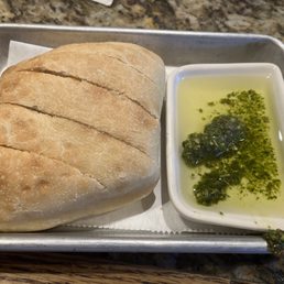 A loaf of bread sliced into sections is served with a small dish of green herb oil or sauce on a metal tray.