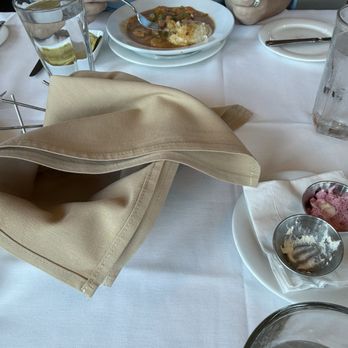 Dauphin's - Shrimp'n Grits - A person wearing a blue shirt and sunglasses hanging from their collar is seated at a restaurant table, eating shrimp and grits with a spoon. The table has a glass of water, a small plate with a spoon, and desserts in small bowls. A beige napkin is draped over the table, and the setting appears formal with a white tablecloth. - Seafood Near Me - Mobile, Alabama
