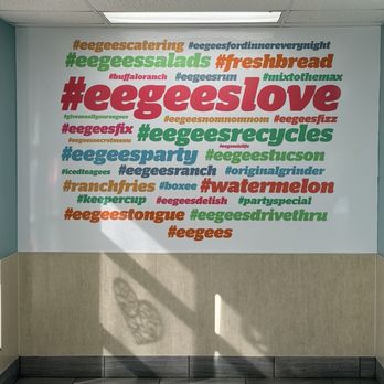 eegee's - The image shows a wall decorated with multiple hashtags and phrases in different colors, such as red, green, blue, and orange, related to Eegee's, a restaurant or food business. The wall has a white background with a beige lower section, and the ceiling has fluorescent lights. Sunlight creates shadows on the lower part of the wall and the floor, including a shadow of a patterned object. The floor is tiled with gray tiles, and the overall setting appears to be indoors, possibly in a restaurant or office space. - Sandwiches Near Me - Tucson, Arizona