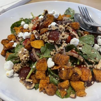 Wildflower - A salad with roasted sweet potatoes, cherry tomatoes, leafy greens, grains, and cheese crumbles, presented on a white plate. The dish includes various textures and colors, with a fork and spoon placed on the side, indicating it is ready to eat. - Sandwiches Near Me - Tempe, Arizona