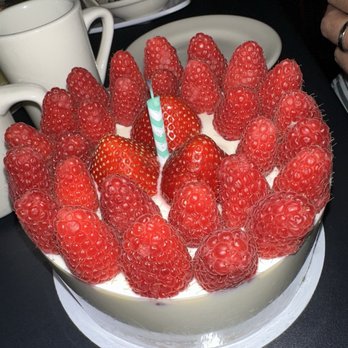 La Saison - A round cake decorated with fresh strawberries and raspberries on top, with a single striped candle inserted among the berries. The cake is on a white cake board, and the background includes cups and bowls with food, indicating a celebratory setting. - Bakeries Near Me - Boston, Massachusetts