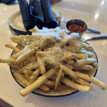 The Kitchen Table - Parmesan garlic fries. - A bowl filled with French fries topped with grated Parmesan cheese and herbs is placed on a restaurant table. The fries are golden and crispy, with cheese sprinkled on top. A small metal container of ketchup is positioned nearby. The table surface is light-colored, and there are utensils, napkins, and a glass of water visible in the background. - American Near Me - Clermont, Kentucky