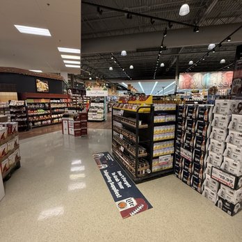Strack & Van Til - Extremely clean - The image depicts an aisle inside a grocery store with multiple shelves stocked with various food and beverage products. The shelves are organized and contain items such as snacks, drinks, and other packaged goods. A promotional sign is placed on the floor, advertising Miller Lite beer. The store has a high, industrial-style ceiling with exposed beams and hanging lights. The lighting is bright, and the aisle extends toward the back of the store, where additional shelves and sections are visible. - Bakeries Near Me - Schererville, Indiana