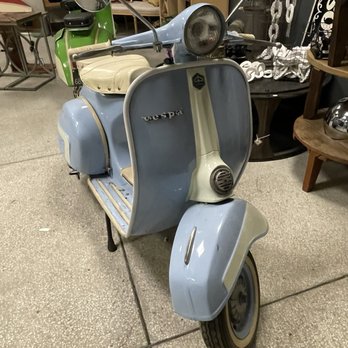 Be Here Now - A baby blue Vespa! - A classic Vespa scooter in light blue with a beige seat is positioned indoors, surrounded by furniture and decorative items. - Furniture Stores Near Me - Independence, Missouri
