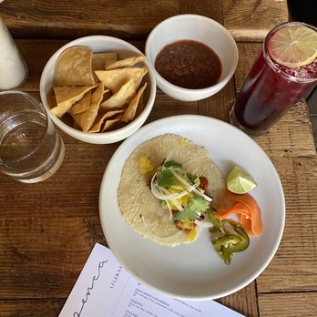 Penca - Diablo cocktail  Al pastor taco - The image shows a wooden table with a white plate containing a taco topped with cilantro, onions, and yellow sauce, along with a lime wedge and pickled jalapeños. Nearby, there is a bowl of chips and a bowl of salsa. To the right, a tall glass of dark red drink with ice and a lemon slice is visible. A glass of water and a menu are also present on the table, indicating a dining setting at a Mexican restaurant. - Mexican Near Me - Tucson, Arizona
