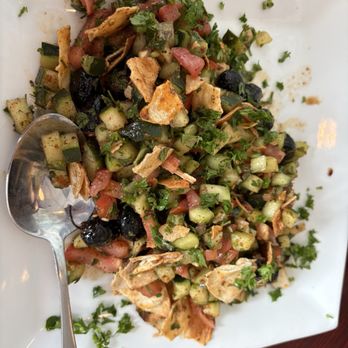 Fettoosh - Fettoosh salad - A chopped salad featuring cucumbers, tomatoes, black olives, and fresh herbs is presented on a white square plate. The salad includes small pieces of pita or flatbread and is garnished with chopped parsley. A metal spoon rests on the plate, and the dish appears to be a Mediterranean or Middle Eastern style salad. - Moroccan Near Me - Arlington, Virginia