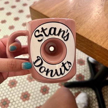 Underground Donut Tour - A person is holding a pink ceramic mug decorated with a donut graphic and the words "Stan's Donuts" in black script. The person's nails are painted light blue, and they are seated near a wooden table. The background shows a patterned floor with small floral tiles and a person's foot wearing a white shoe. The image captures a casual setting, likely in a cafe or donut shop. - Food Tours Near Me - Chicago, Illinois