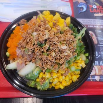 Poke Land Extra - A poke bowl containing vegetables, corn, and crispy toppings, served on a black bowl on a red tray at a restaurant. - Poke Near Me - Redding, California