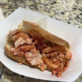 Angie's - Lobster Roll with extra lobster - A lobster roll with chopped lobster meat inside a toasted bun, served on a white paper tray. The lobster meat appears fresh and is mixed with a light sauce. The tray is placed on a speckled granite countertop, and the image captures the food in a casual dining setting. - Chicken Shop Near Me - Tucson, Arizona