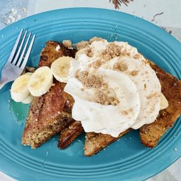 Gluten-free banana bread French toast - A serving of French toast topped with whipped cream, banana slices, and crumbles is presented on a blue plate. The toast appears golden brown, and the toppings include sliced bananas and a dollop of whipped cream. A fork is placed on the left side of the plate, and the background includes a light-colored table surface with some decorative elements.