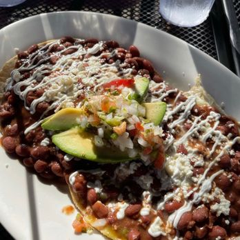 Ray's Luncheonette - Huevos rancheros - A plate of huevos rancheros features beans, cheese, and sauce, garnished with sliced avocado and a mixture of chopped onions, tomatoes, and peppers. The dish is served on a white oval plate, with condiments and glasses of water visible on a black metal table. The setting appears to be a casual dining environment at Ray's Luncheonette in Montclair, NJ. - Breakfast & Brunch Near Me - Montclair, New Jersey