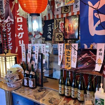 Dragon Sushi - Greeting counter. - A restaurant counter decorated with Japanese-themed items, including a large red paper lantern hanging from the ceiling, banners with Japanese writing, and bottles of sake lined up on the counter. The background features various posters and signs, some with Japanese characters, and a framed photo or artwork. There is a small figurine resembling Baby Yoda on the counter, along with a bottle of hand sanitizer and a transparent container with toothpicks. The overall setting suggests a Japanese restaurant or sushi bar with traditional and decorative elements. - Sushi Bars Near Me - Albuquerque, New Mexico
