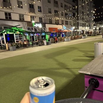 Killer Queen - High Noon - Nighttime outdoor scene at a commercial area with string lights hanging overhead. The area features storefronts with illuminated signs, outdoor seating, and a few people walking or sitting. The foreground shows a person holding an open can, with a table and a purple structure nearby. The background includes a multi-story building with windows and balconies. - Pizza Near Me - Cincinnati, Ohio