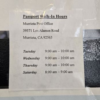 US Post Office - Passport Walk-In Hours as of 9.10.2024 - The image features a printed sign with the title 'Passport Walk-In Hours' for the Murrieta Post Office, located at 39571 Los Alamos Road, Murrieta, CA 92563. The sign details specific days and times for passport walk-in services: Tuesday, Wednesday, and Thursday from 9:00 am to 10:00 am, and Saturday from 8:00 am to 9:00 am. The sign is taped to a surface, with some reflections visible on the plastic covering. - Post Offices Near Me - Murrieta, California