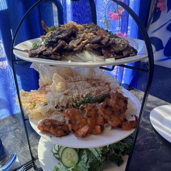 Dan Da - RP2. Tu Quy Tower - A three-tiered serving stand displays Vietnamese cuisine with beef, fried items, and fresh vegetables, set against a blue curtain background. - Vietnamese Near Me - Aurora, Colorado