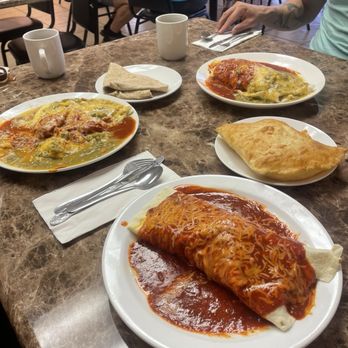 Ramblin'cafe - The image shows a restaurant table with three plates of Mexican food, including two enchiladas covered in sauce and cheese, and a calzone-style dish. There are also chips on a small plate, two coffee mugs, and utensils on a napkin. Diners are seated in the background, and the setting appears casual with a marble-like table surface. - Kosher Near Me - Santa Fe, New Mexico