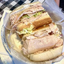 Trans-Am Turkey Sandwich - A sandwich featuring sliced turkey meat and lettuce on a hoagie bun, presented in a foil tray with parchment paper. The sandwich appears to be freshly made, with visible layers of turkey and crisp lettuce. The background includes a checkered tablecloth, indicating a casual dining setting.