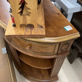 Savers - Beautiful oak furniture! - A brown wooden corner cabinet features a curved front with a single drawer at the top and an open shelf below. The cabinet has decorative vertical grooves on the sides and a round wooden knob on the drawer. It is placed on a beige tiled floor, with other furniture and items visible nearby, including a white shelf and a decorative wooden piece with a Christmas tree cutout. - Thrift Stores Near Me - Vacaville, California