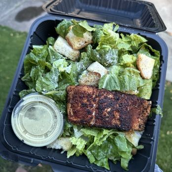 Charred - Caesar salad with blackened salmon - The image shows a takeout salad in a black plastic container, containing chopped lettuce, croutons, a piece of blackened salmon, and a small cup of dressing. The container is open, revealing the fresh greens and crispy croutons, with the salmon placed on top. The background suggests an outdoor setting with a sidewalk and grass. - Burgers Near Me - Pittsburgh, Pennsylvania