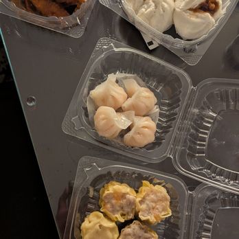 39 Prince Cafe - Siu mai, shrimp dumplings, cha siu baos, chicken feet - Multiple plastic containers hold different dim sum dishes on a gray table. One container has siu mai, another has shrimp dumplings, and a third contains cha siu baos. The dishes are typical of Hong Kong-style late-night dim sum offerings. - Dim Sum Near Me - Flushing, New York