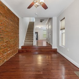Before painting brick wall - The image shows an interior space with hardwood flooring and a brick accent wall on the left side. White walls and trim surround the room, with two windows on the right side allowing natural light. A staircase with beige carpet ascends to the upper level, with a small step down into the main area. Ceiling fixtures provide additional lighting, and the space appears to be part of a residential home.
