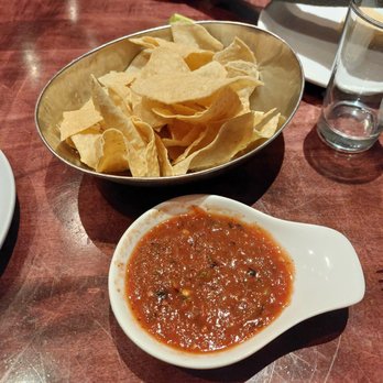 Rosa Mexicano - Complimentary Chips and Salsa - A metal bowl filled with tortilla chips and a small white dish of salsa on a wooden table, with a glass of water nearby. - Mexican Near Me - Hackensack, New Jersey