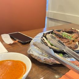 Lamb Chops - Lamb chops with green herbs are placed on a foil-covered tray, with tongs resting on top. A bowl of red sauce is nearby, and a smartphone is on the table.