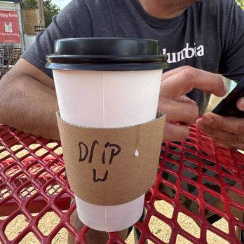 Streamway Coffee - Regular drip coffee - A person is sitting at a red metal outdoor table, wearing a dark T-shirt and looking at a smartphone. The coffee cup in front of them has a black lid, a white sleeve with "DRIP" written on it, and is placed on the table. The background shows outdoor elements such as trees and a sign, indicating an outdoor seating area. - Coffee & Tea Near Me - Austin, Texas