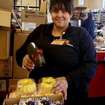 Food King - Mariah (helpful employee) - A woman in a Food King uniform scans items at a grocery store checkout counter, with shelves and other shoppers in the background. - Grocery Near Me - Albuquerque, New Mexico