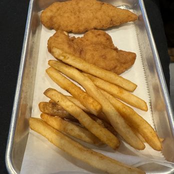 Wolfies Grill - Pendleton - Kids chicken tenders and fries - The image shows a metal tray lined with white paper, holding a fried fish fillet, a chicken tender, and a portion of French fries. The food items are arranged with the fish fillet at the top, the chicken tender below it, and the fries occupying the lower part of the tray. The tray is placed on a dark surface. - Bars Near Me - Pendleton, Indiana