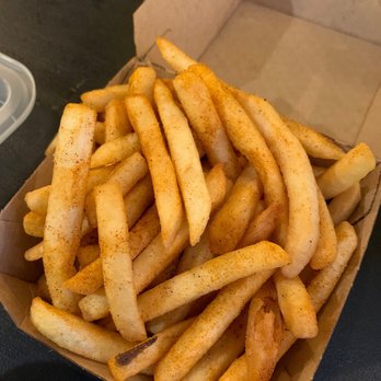 Filly Luv - Classic Luv Fries with cajun seasoning - French fries seasoned with Cajun spices are shown in a small, open cardboard container. The fries are golden and slightly crispy, with visible seasoning on their surface. The container is placed on a dark table or countertop, with part of a beige wall or partition visible in the background. The image captures a casual food presentation typical of a fast-food or snack setting. - Chicken Shop Near Me - Brooklyn, New York