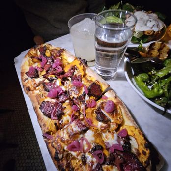 The Binc - Short rib flat bread - A rectangular pizza with various meats and pickled onions, served with a glass of water, a cocktail, and side dishes including salad and grilled vegetables. - Cocktail Bars Near Me - Brooklyn, New York