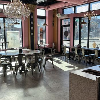 Voodoo Doughnut - Interior - The image shows the interior of a cafe with pink walls and large windows allowing natural light to fill the space. There are two chandeliers hanging from the ceiling, along with several pendant lights. Metal chairs are arranged around round and rectangular tables. The floor appears to be made of light-colored tiles. An exit sign is visible above a door, and the overall decor includes ornate details and a mix of vintage and modern elements. - Donuts Near Me - Denver, Colorado