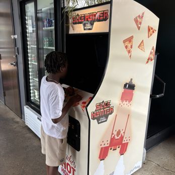 Futuro - Space shooter video game - A child with dark, curly hair, wearing a white t-shirt, beige shorts, and sneakers, is standing in front of an arcade game called 'Slice Hunter.' The game features a pizza-themed design with pixel art of a giraffe-like creature and pizza slices on the side. The setting appears to be indoors, with a refrigerator stocked with beverages visible nearby and some potted plants on top of the arcade machine. - Pizza Near Me - Indianapolis, Indiana