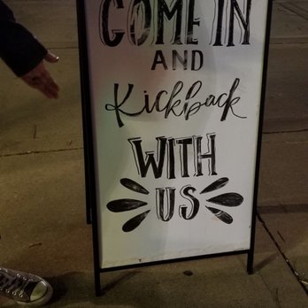 Kickback Cafe - come in and kickback with us sign - A freestanding sidewalk sign with a white background and black lettering reads "Come in and kick back with us" in different font styles. The sign is positioned on a sidewalk in an urban area during nighttime, with streetlights, buildings, and parked cars visible in the background. Part of a person's hand and shoe are visible on the left side of the image. - Cafes Near Me - Oregon, Wisconsin