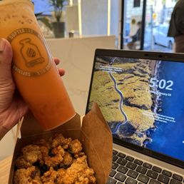 Popcorn Chicken - The image shows a person holding a cup of orange-colored drink with a logo on it, along with a box of popcorn chicken on a table. A laptop displaying a scenic landscape is also on the table. The cafe interior features a white counter, a potted plant, and a partially open door with a view of the street outside.