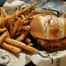 Buffalo Chicken Sandwich - A chicken sandwich featuring a fried chicken fillet inside a soft bun, secured with a toothpick, is placed on a black tray lined with printed paper. Next to the sandwich, there is a portion of French fries, some of which are seasoned or lightly salted. The setting appears to be a casual dining environment, possibly a bar or tavern, with a dark wooden table and a glass of water nearby.