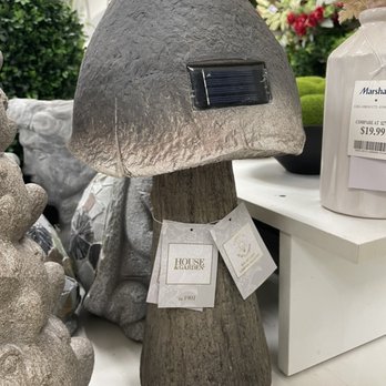 Marshalls - Cool solar powered mushroom garden light - A mushroom-shaped garden light features a textured, gray cap with a small solar panel attached to its front surface. The stem is designed to resemble natural wood or bark, providing a rustic appearance. The light is displayed on a white surface in a retail store, surrounded by other decorative items and greenery. A tag hanging from the stem indicates it is part of the House & Garden collection. - Department Stores Near Me - Costa Mesa, California