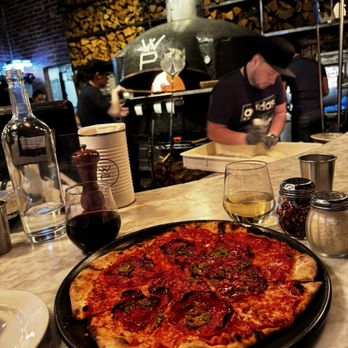White Pie - A pizza topped with jalapeños and pepperoni on a black plate, with drinks and condiments on a marble table in a restaurant setting. - Pizza Near Me - Denver, Colorado
