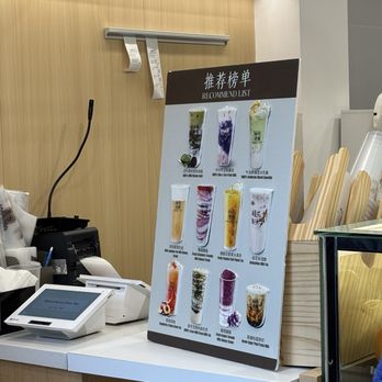 Choo Tea - Counter inside - A menu board featuring images and names of different bubble tea drinks, positioned on a white counter in a tea shop. The counter has a point-of-sale system and some supplies nearby. The background includes a wooden wall with a metal hook and receipt holder, and a basket with paper straws. - Bubble Tea Near Me - Toronto, Ontario