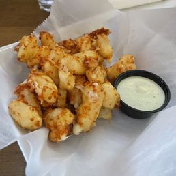 Wisco Cheese Curds - A basket contains fried cheese curds, which are golden-brown and crispy on the outside. The cheese curds are placed on white parchment paper, with a small black container of creamy dipping sauce beside them. The background shows a glass of water and a napkin on a wooden table, indicating a casual dining setting.
