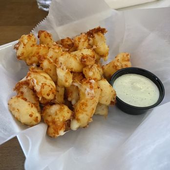 Commonwealth - Wisco Cheese Curds - A basket contains fried cheese curds, which are golden-brown and crispy on the outside. The cheese curds are placed on white parchment paper, with a small black container of creamy dipping sauce beside them. The background shows a glass of water and a napkin on a wooden table, indicating a casual dining setting. - American Near Me - Chicago, Illinois