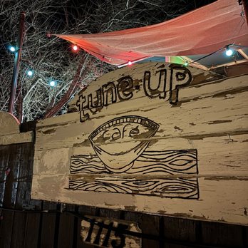 Tune-Up Cafe - The image shows a rustic wooden sign for Tune-Up Cafe, featuring hand-painted text and a coffee cup illustration on a distressed, white-painted surface. The sign is mounted on a fence or wall, with a small additional sign displaying the number 715. Overhead, colorful string lights are strung across the scene, and a pink canopy or fabric is visible at the top, with leafless trees in the background indicating a nighttime setting. - New Mexican Cuisine Near Me - Santa Fe, New Mexico