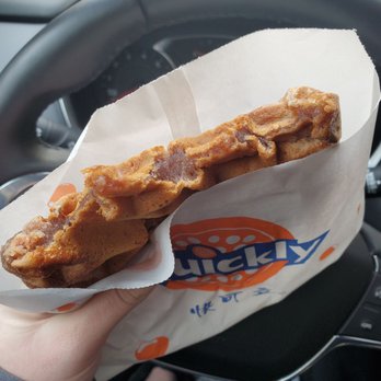 Quickly - Ann Arbor - Nutella stuffed waffle. Yum! - A hand holds a Nutella stuffed waffle wrapped in a paper bag with a logo, inside a car. The waffle appears golden brown with Nutella filling visible on the surface. The background shows the steering wheel and dashboard of the vehicle, indicating the photo was taken from the driver's seat. - Bubble Tea Near Me - Ann Arbor, Michigan