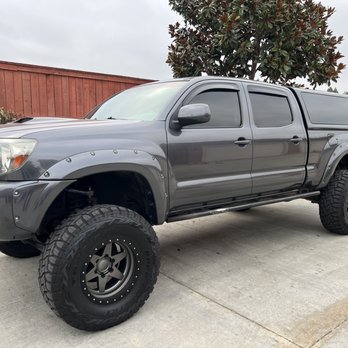Carfection Pro - Body straight and good clear applied. - A gray pickup truck with custom modifications, including large off-road tires, black rims, and riveted fender flares, is parked on a concrete surface. The vehicle has a canopy over the bed and tinted windows. It is positioned near a wooden fence with trees in the background, and the sky appears overcast. - Body Shops Near Me - Oxnard, California