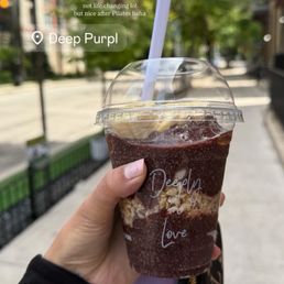 Acai Bowl - A person holds a plastic cup with a dark beverage and a straw outdoors, with trees and buildings visible in the background.