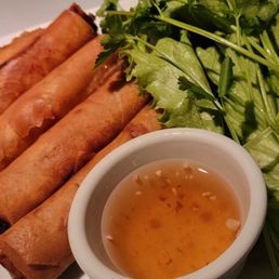 Egg Roll - Five spring rolls are arranged on a white plate with a small bowl of dipping sauce and fresh greens, likely served at a Vietnamese restaurant.